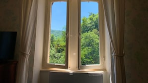 A window with white curtains, a vent below, and a view of green trees and a clear sky.