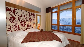 A hotel room with a large bed, a decorative headboard, a nightstand, a lamp, and a window with a mountain view. - Apartment in Les Arcs Near Paradiski Slopes (Bourg-Saint-Maurice)