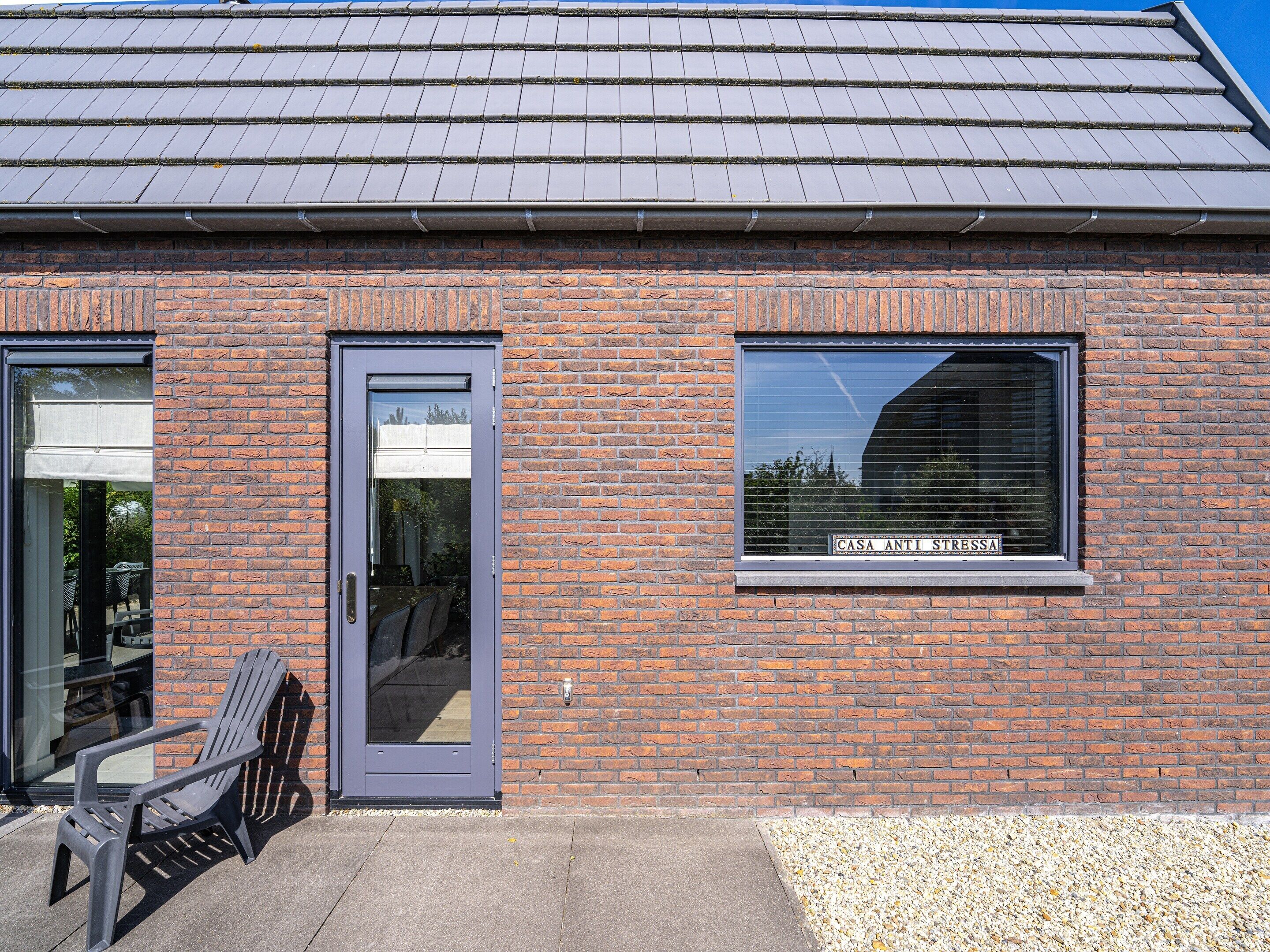 A brick building with a grey door and window, a patio with a chair, and a sign reading "CASA ANTI STRESSSA".