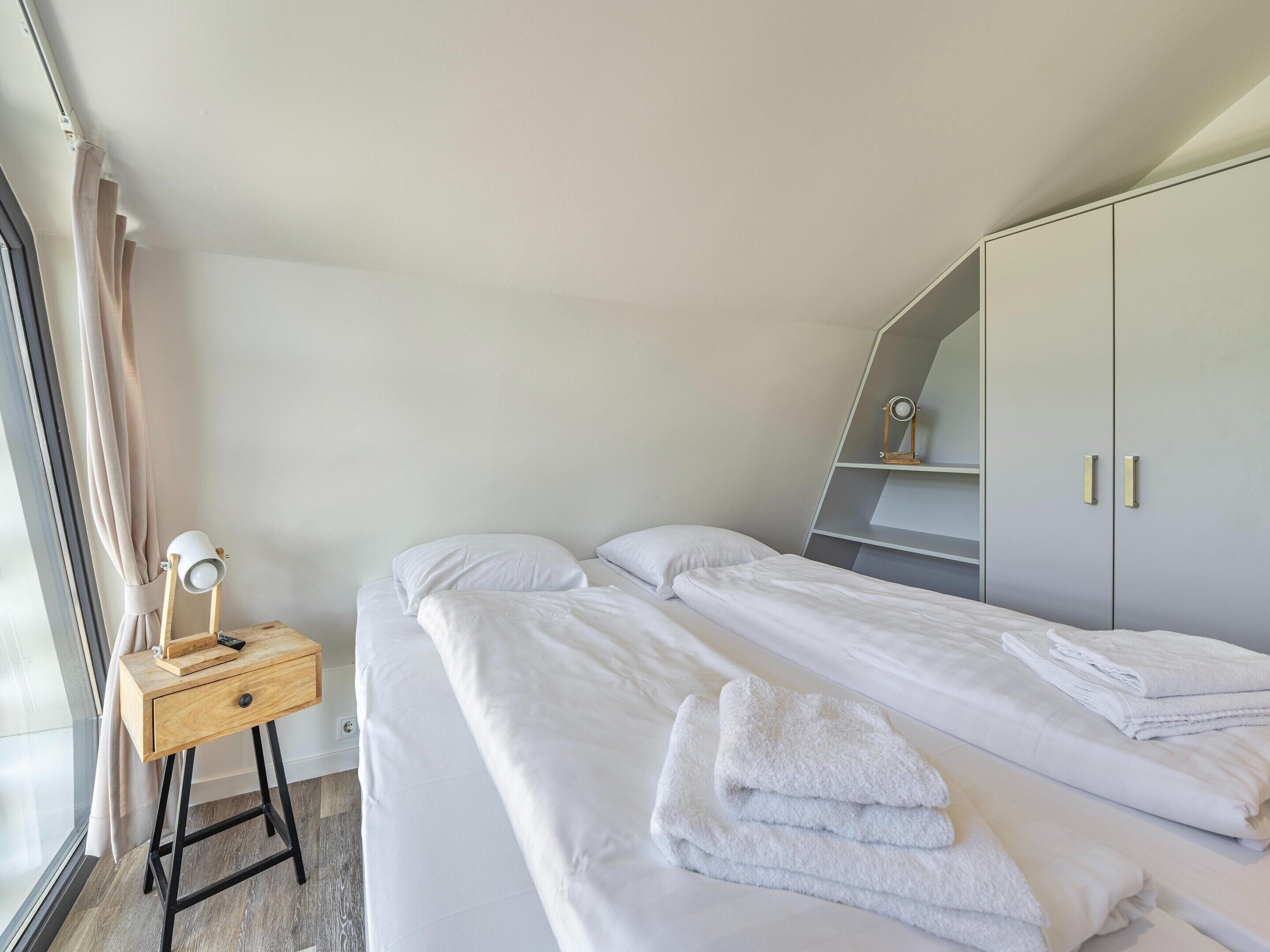 A bedroom with two beds, a bedside table with a lamp, a wardrobe, and a window with curtains.
