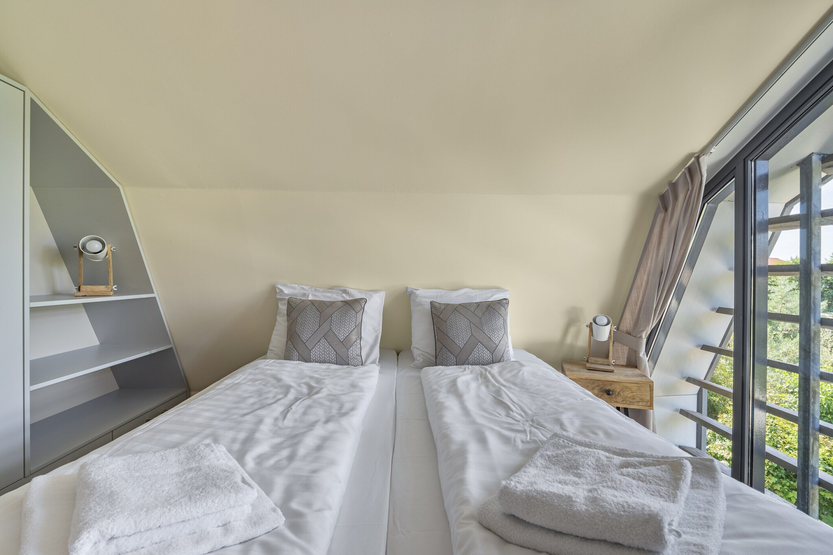 Une chambre avec deux lits, une table de chevet en bois, une étagère et une fenêtre donnant sur un paysage verdoyant.