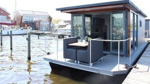 Une maison flottante moderne dotée d’un espace repas, de grandes fenêtres et d’un bateau amarré en arrière-plan.