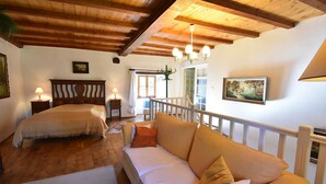 Un dormitorio con cama, sofá, vigas de madera, candelabro, ventana y un cuadro.