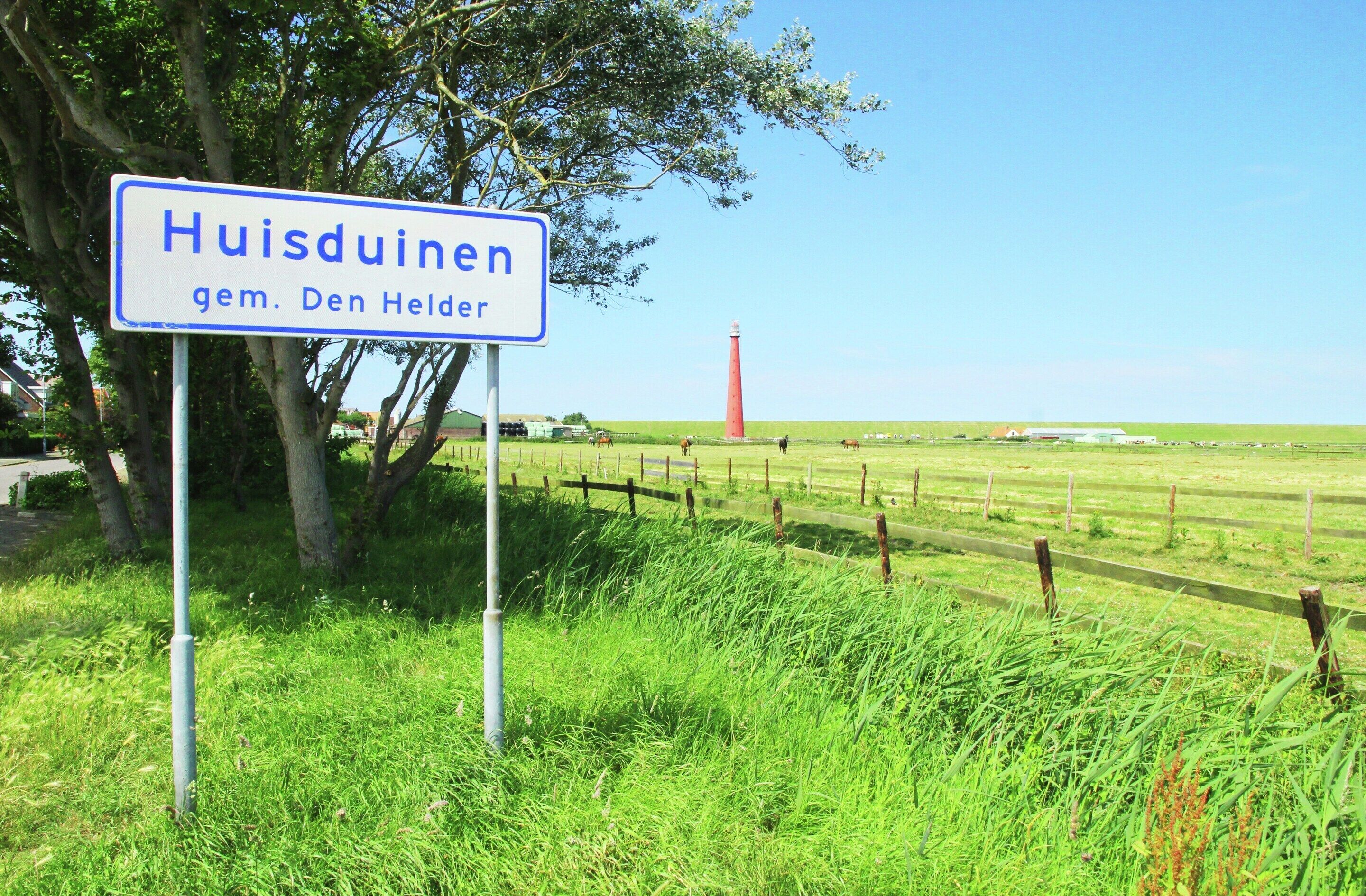 A sign for Huisduinen gem. Den Helder with a red lighthouse in the background.