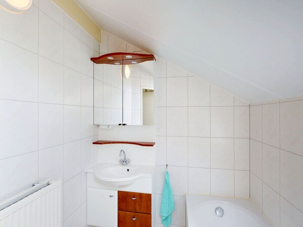 A bathroom with white tiles, a white sink, a wooden vanity, a mirror, and a towel hanging on the wall.