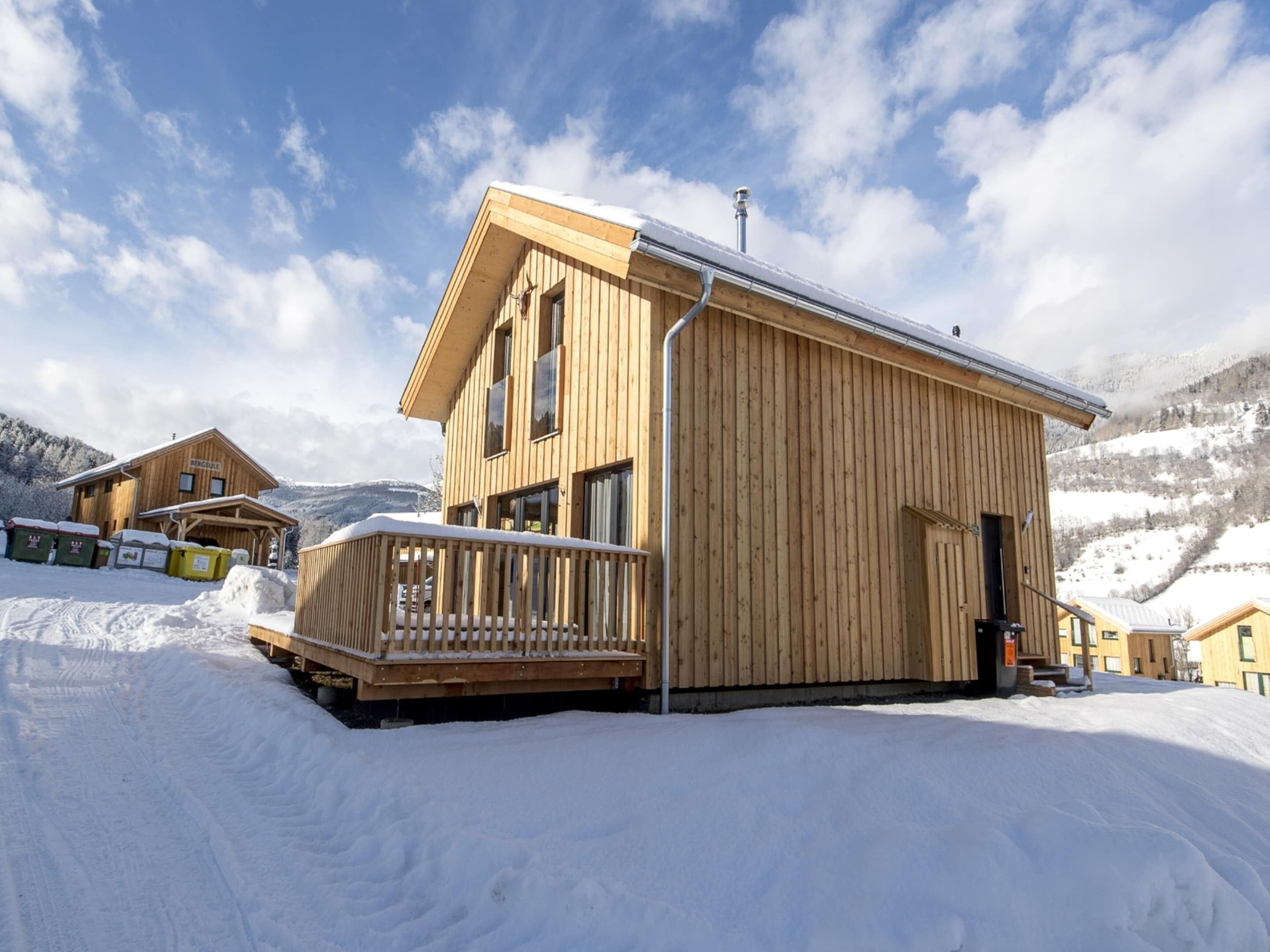 A wooden chalet with a balcony, a covered entrance, and a view of snow-covered mountains.