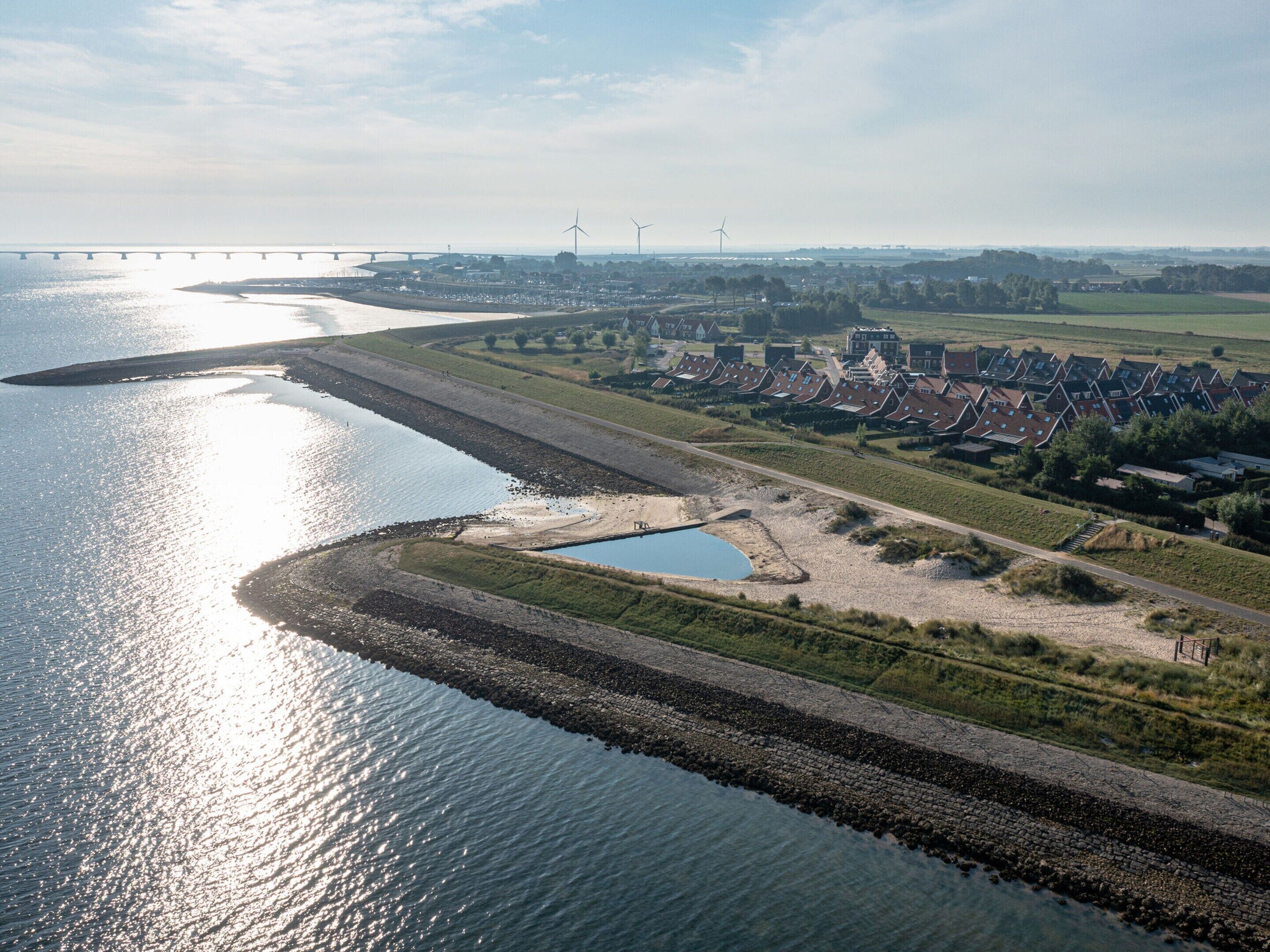 An aerial view of a riverside town with a bridge in the background, a small lake, and wind turbines.