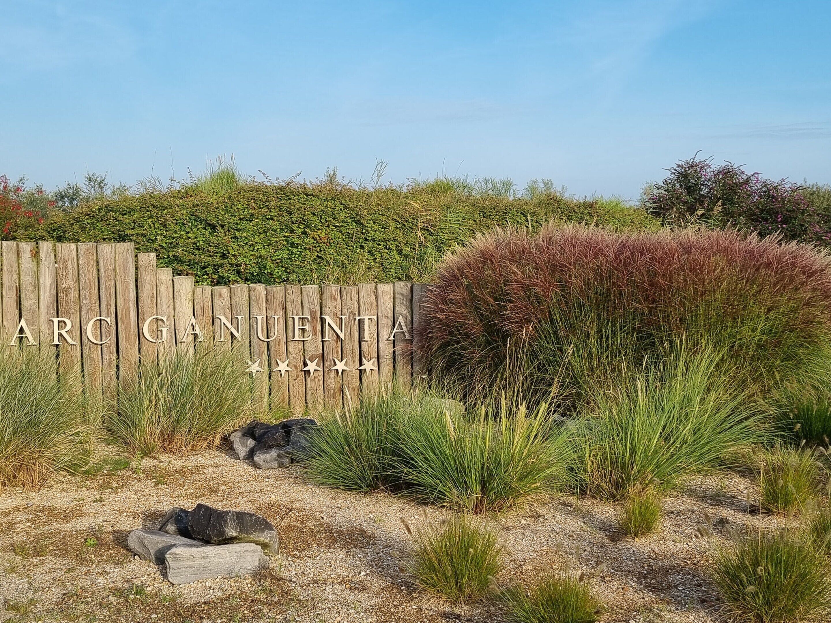 Een houten hek met de tekst 'ARC GUENTA' en een aangelegde tuin met diverse planten en rotsen.