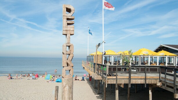 Une jetée au bord de la plage, ornée d’un drapeau de Texel, dotée d’une terrasse en bois et de parasols jaunes.