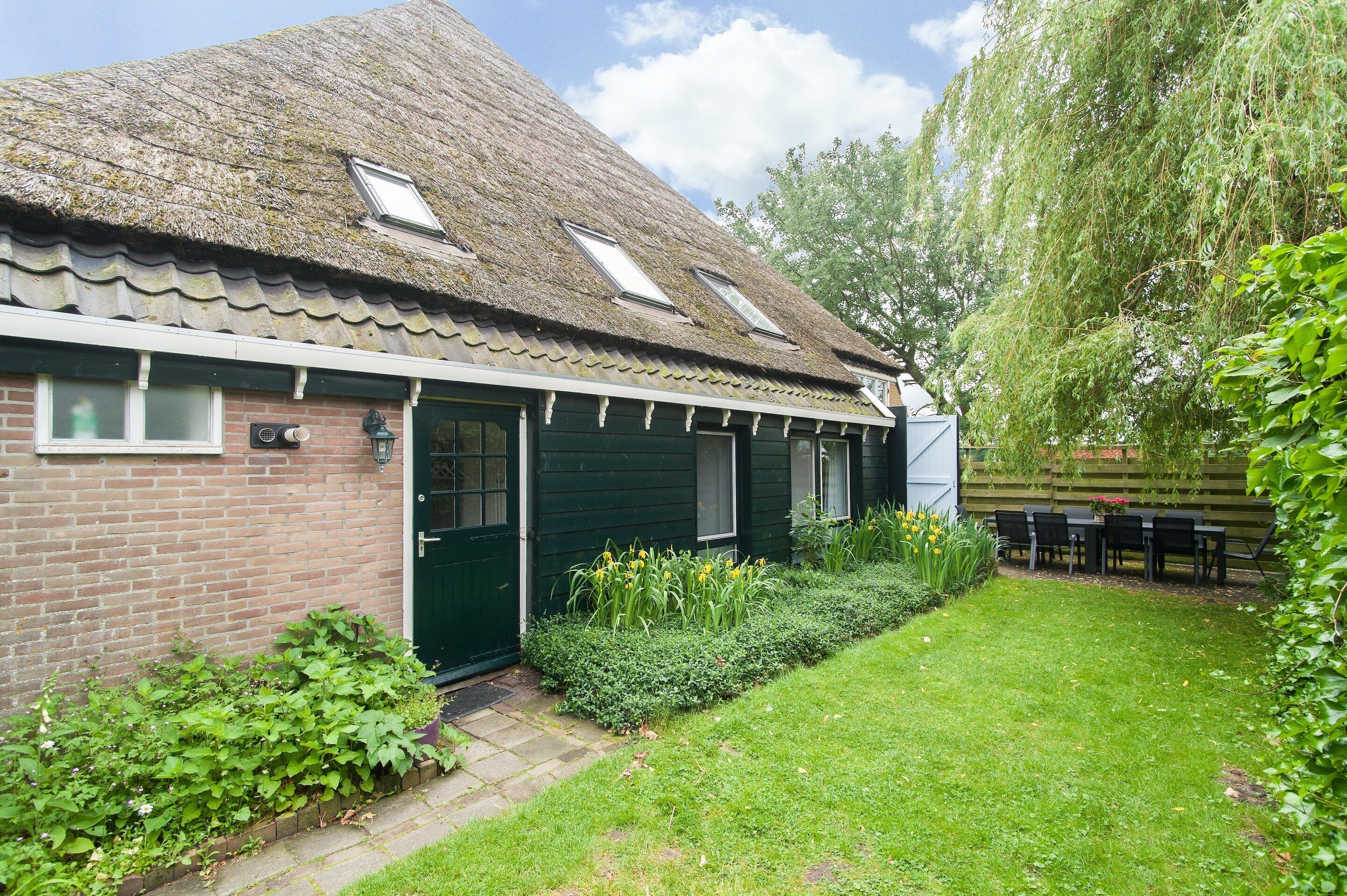 A traditional house with a sloped roof, a green door, and a small garden with a lawn and a table with chairs.