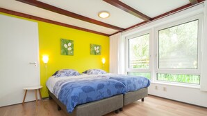 Une chambre à coucher comprenant un lit, une petite table, deux lampes fixées au mur et de grandes fenêtres donnant sur un paysage verdoyant.