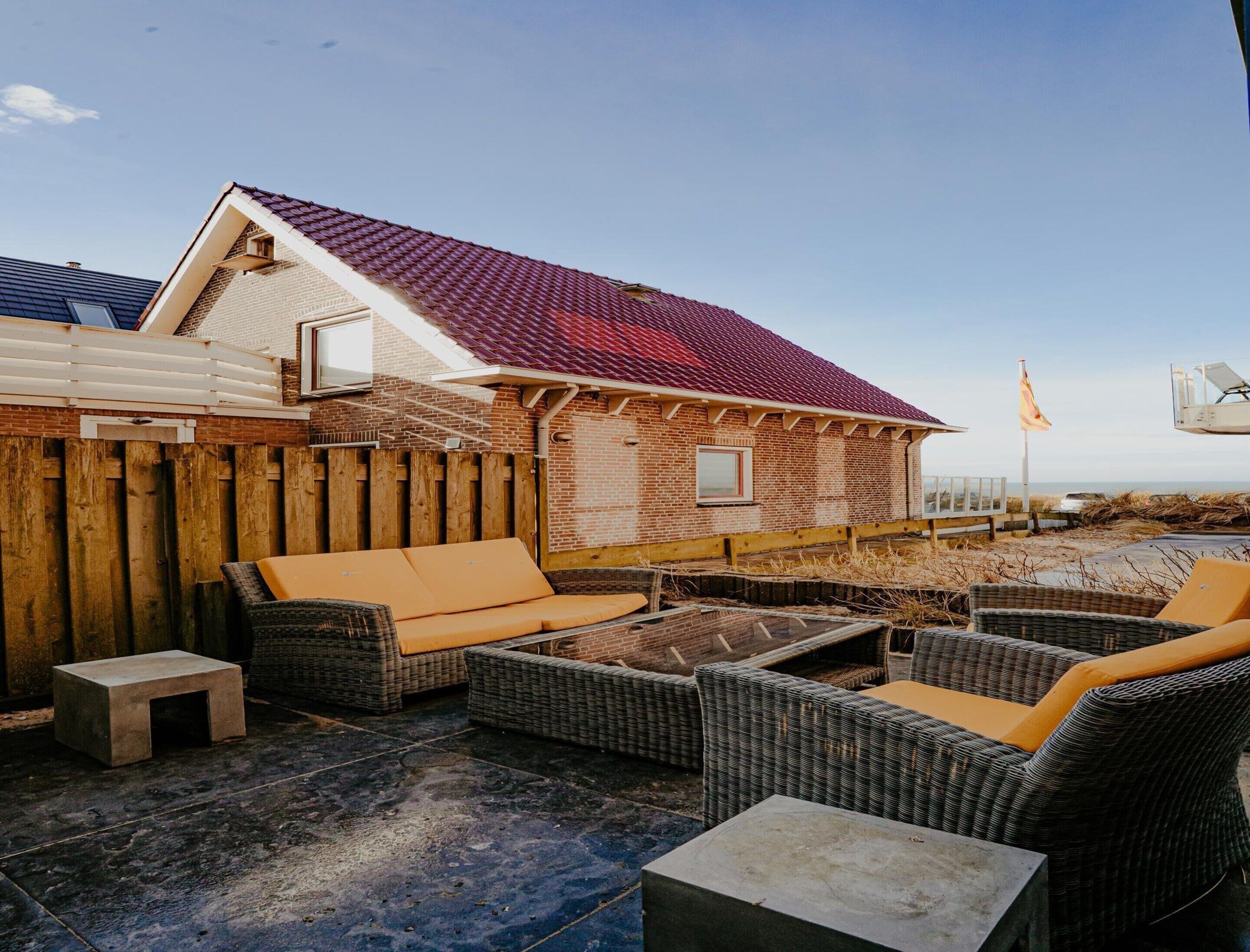 A patio area with wicker furniture and a small concrete table, a brick house with a red roof, and a wooden fence.