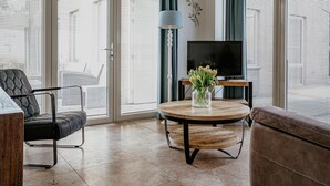 A modern living room with a sofa, a chair, a coffee table with a vase of flowers, a TV on a stand, and large windows with blinds. - Apartment in Egmond With Stunning Sea Views (Egmond aan Zee)