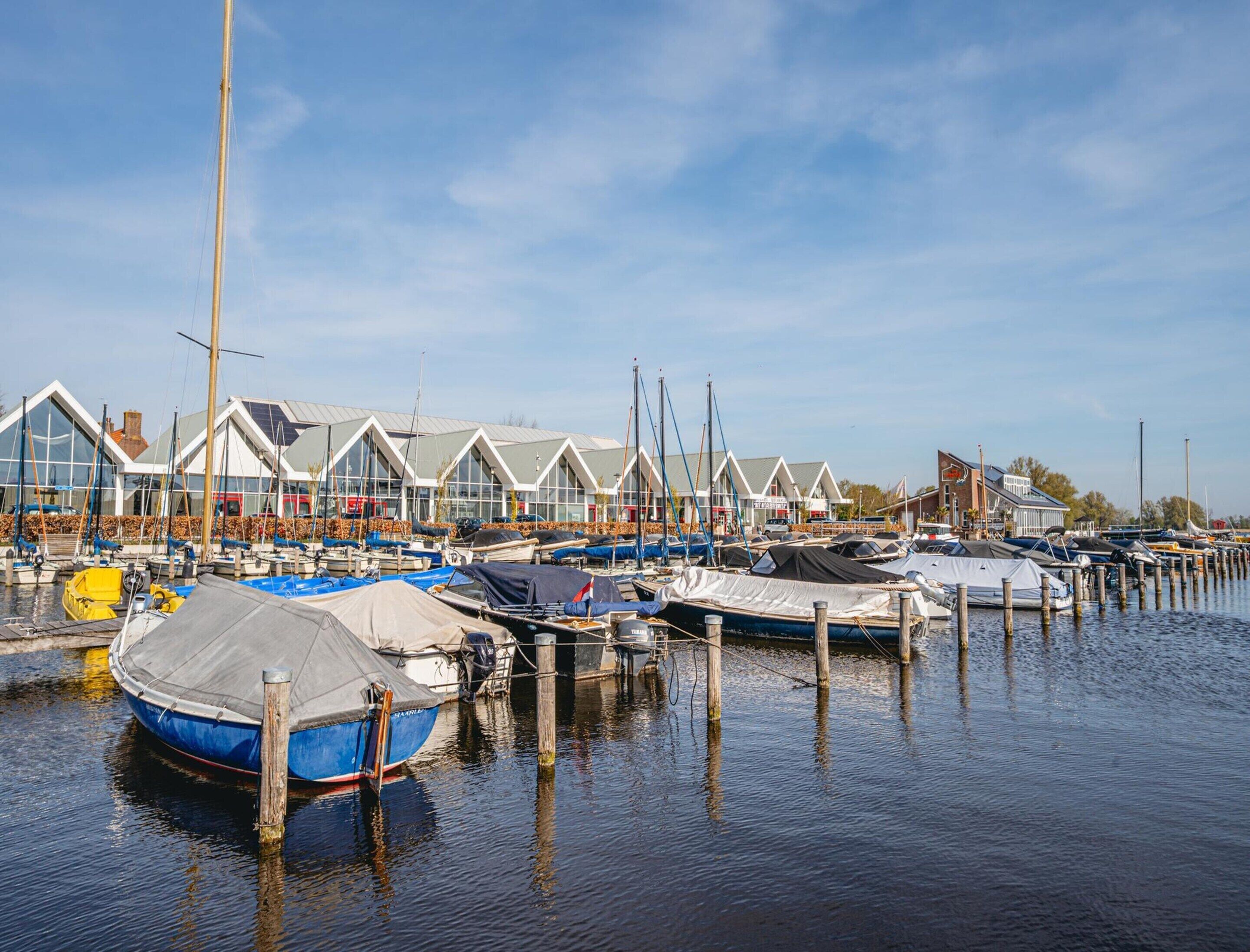 A marina with boats docked at wooden piers, a row of modern buildings with large windows, and a clear sky.