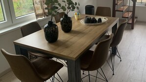 A dining area with a wooden table, four chairs, two vases with plants, a tray with cups, a hanging light fixture, and a staircase.