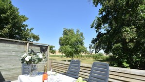 An outdoor dining area with a table set for two, featuring a bottle of wine, glasses, and a bowl of apples.