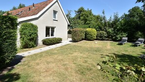 A house with a tiled roof, a front door, and a small window. There is a paved path leading to the door, and a lawn with a few plants and a bench.