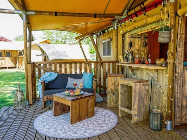 Una terraza de madera al aire libre con una mesa redonda, un sofá con cojines y una zona de bar con botellas y fruta.