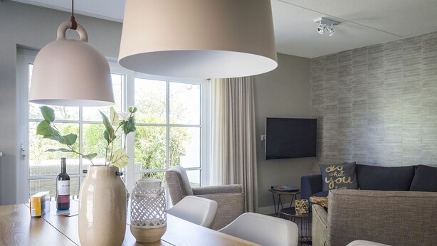 A modern living room with a wooden dining table, two hanging lamps, a large window with curtains, a flat-screen TV mounted on the wall, and a sofa with decorative pillows. - Apartment in De Cocksdorp With Garden (De Cocksdorp)