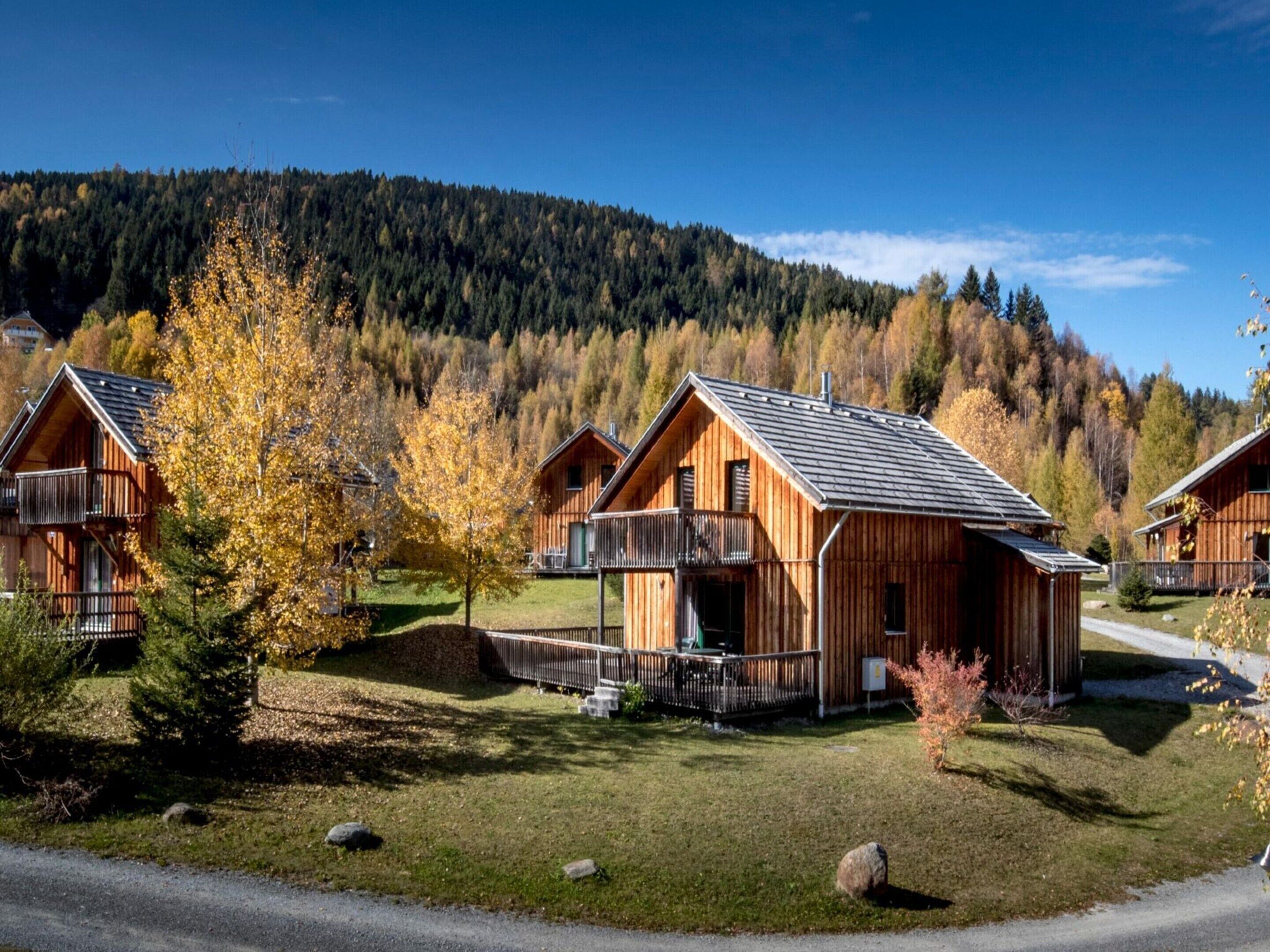 A row of wooden cabins with balconies, set against a backdrop of autumn-colored trees and a mountain.
