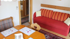 A cozy living room with a red sofa, wooden headboard, and a dining table with floral placemats. - Apartment in Nassfeld (Hermagor-Pressegger See)