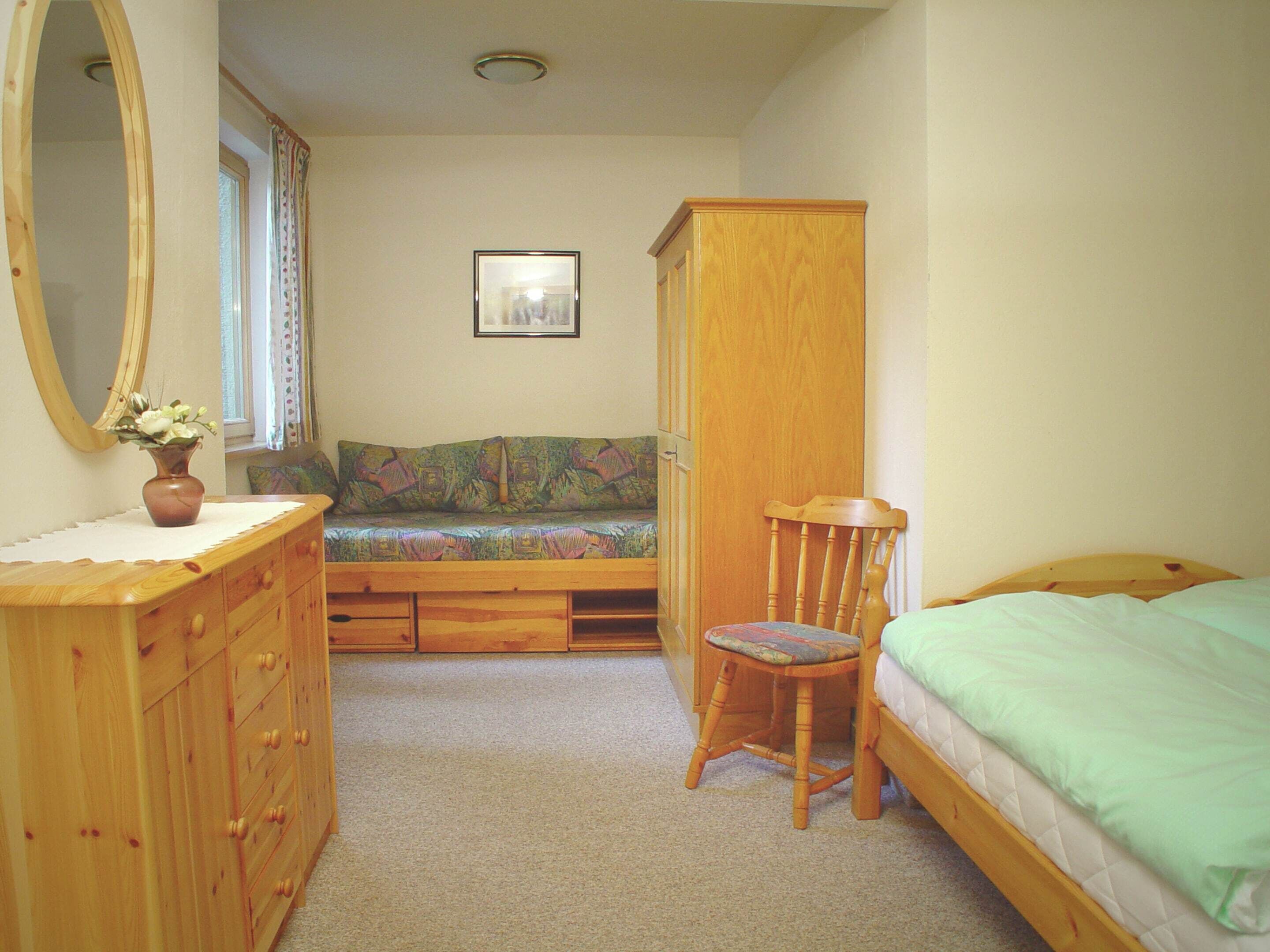 A bedroom with a wooden dresser, a chair, a bed, and a window with curtains.