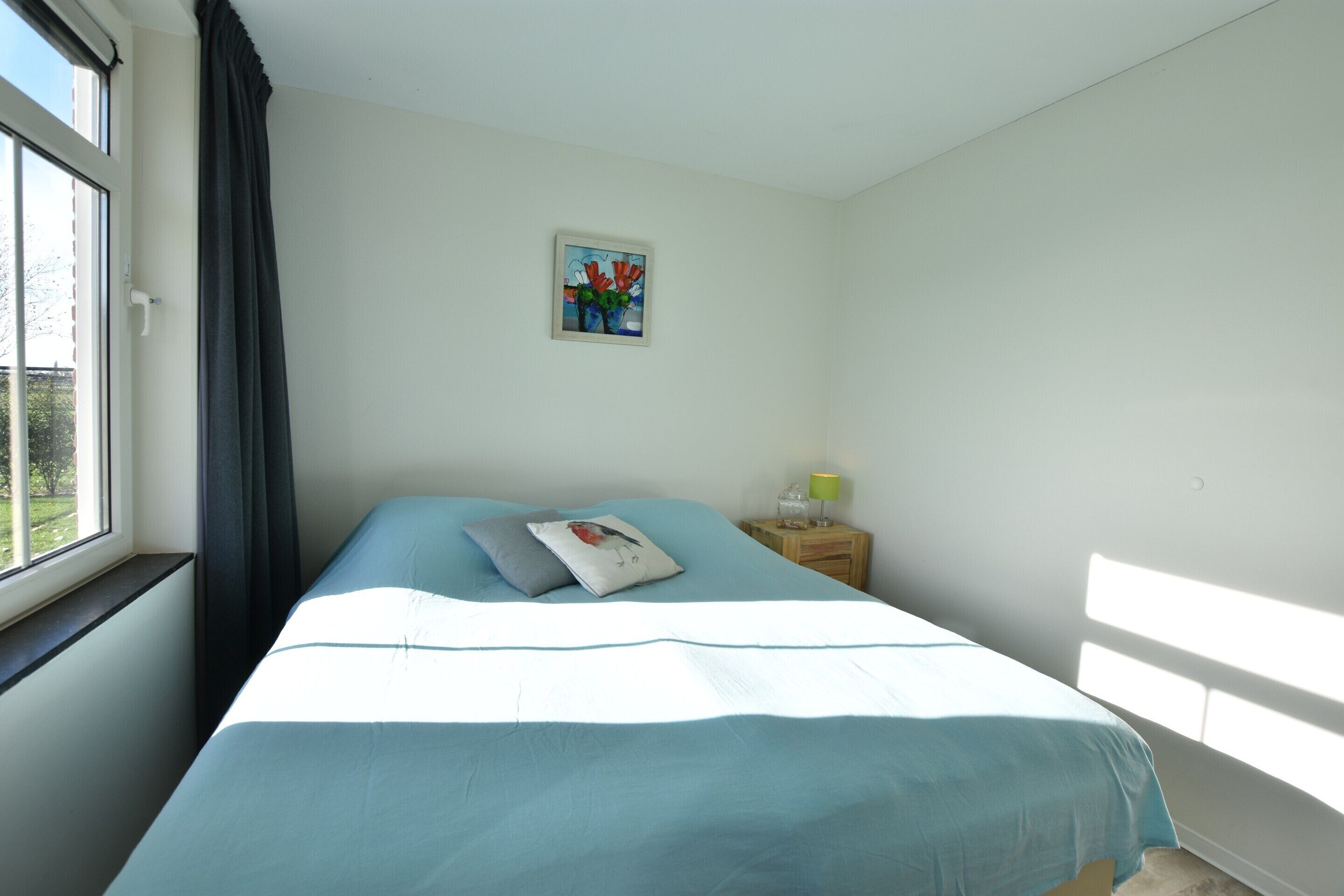 Ein Schlafzimmer mit Bett, Nachttisch, einem Fenster mit Vorhängen und einem Wandbild.