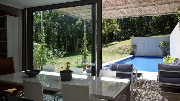 A modern outdoor dining area with a glass table, white chairs, and a view of a swimming pool and garden. - Holiday Home in Boissieres With Private Pool (Boissieres)