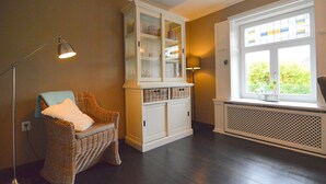 Una habitación con una silla de mimbre, un armario blanco con puertas de cristal, una lámpara de pie y una ventana con vistas a zonas verdes.
