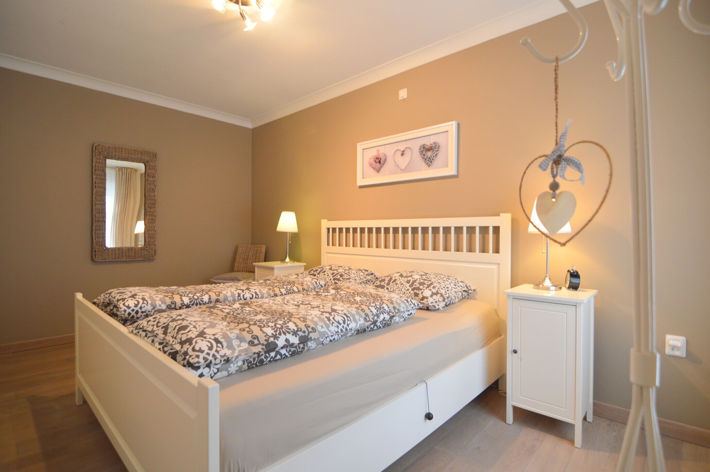 A bedroom with a white bed, a nightstand with a lamp, a small cabinet, and wall decorations including a heart-shaped hanging ornament.