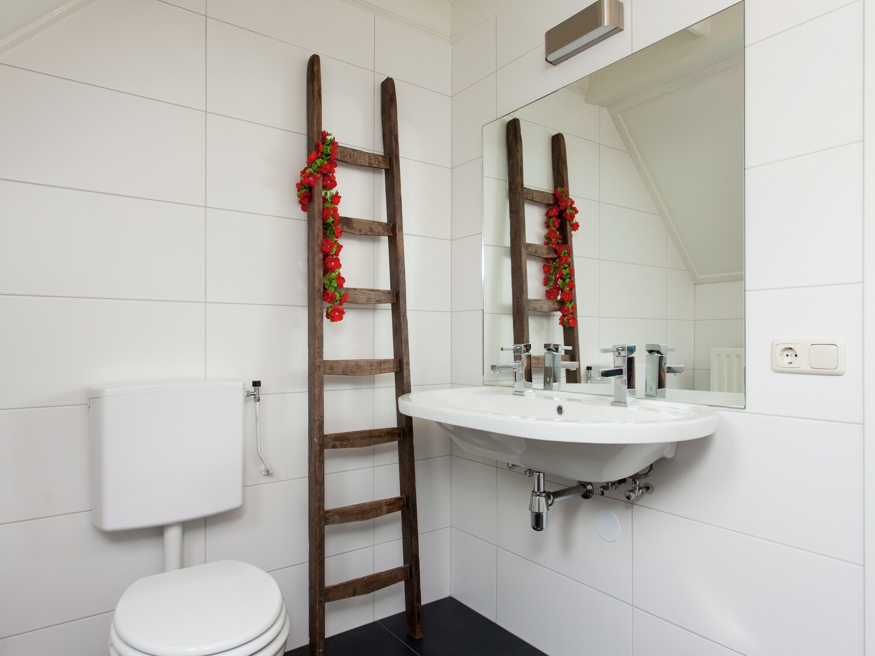 Un baño con inodoro y lavamanos blancos, y un espejo sobre el lavamanos. Se ve una escalera de madera apoyada en la pared con flores rojas.