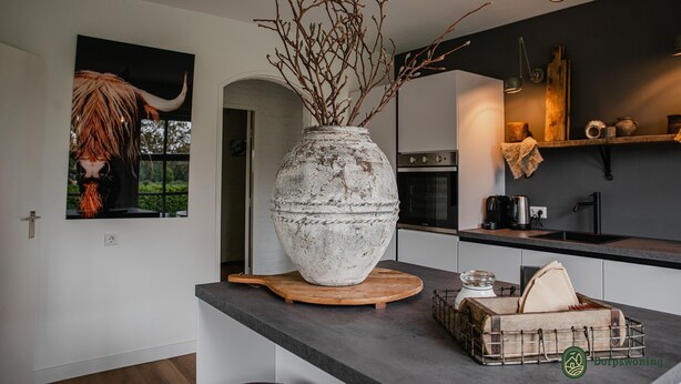 A kitchen with a large white vase with branches, a wooden cutting board, and a metal basket on the counter. - Forest Cottage Near Leenderbos (Leende)
