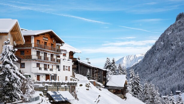 A snow-covered mountain resort with multi-story buildings, ski slopes, and a forested backdrop.