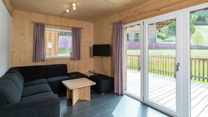 Une pièce chaleureuse avec un plafond en bois, un canapé noir, une petite table en bois et un téléviseur. Une porte-fenêtre coulissante donne accès à une terrasse extérieure.