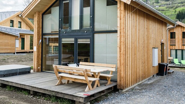 Una baita di legno con un'ampia porta vetrata, un terrazzino in legno con una panchina e vista su un paesaggio collinare.