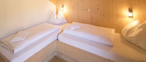 Una habitación pequeña y acogedora con dos camas de madera, cada una con ropa blanca y almohadas, y dos lámparas fijadas a la pared.