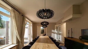 Uma longa mesa de jantar de madeira com cadeiras, um lustre preto pendurado e uma televisão em um armário.