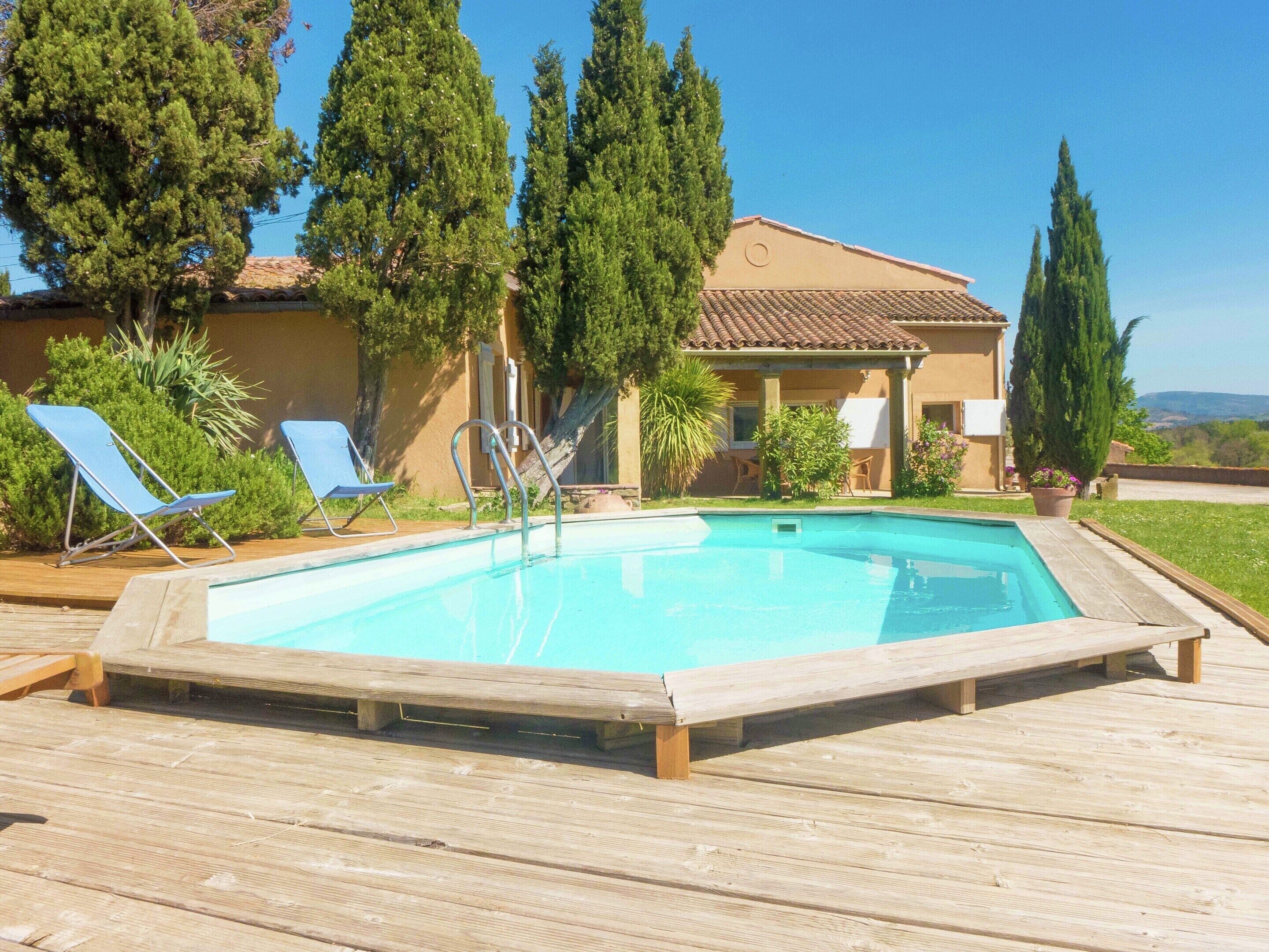 A swimming pool with a wooden deck, lounge chairs, and a building with a tiled roof.