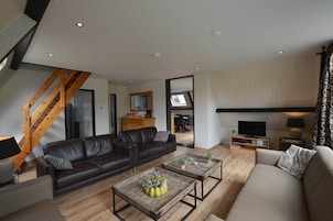 Una sala de estar moderna con un sofá de cuero marrón, un sofá beige y dos mesas de centro con objetos decorativos. Se observa una escalera de madera al fondo.