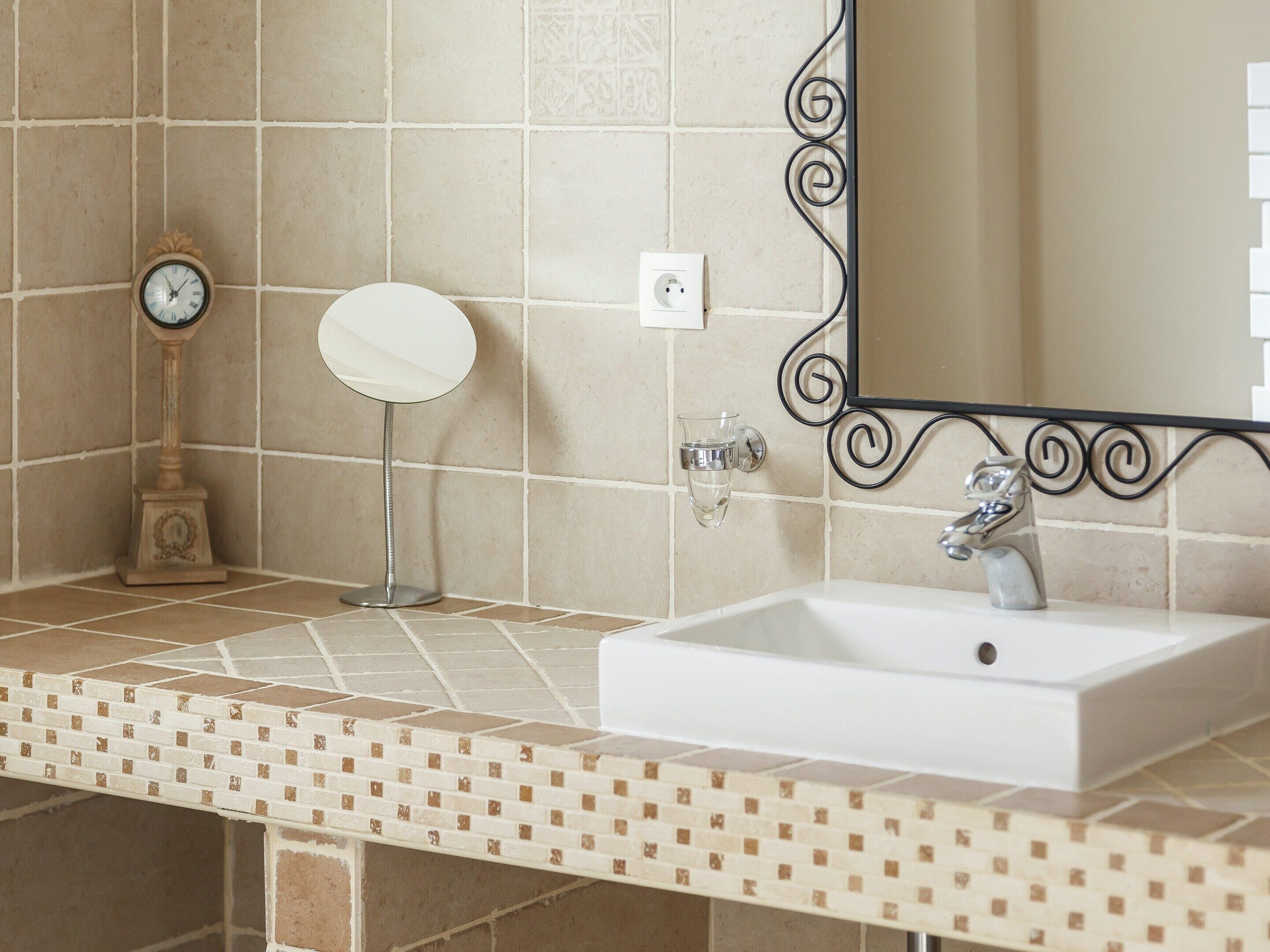 Une salle de bain avec un lavabo blanc, un miroir doté d’un cadre décoratif, une horloge murale et un interrupteur.