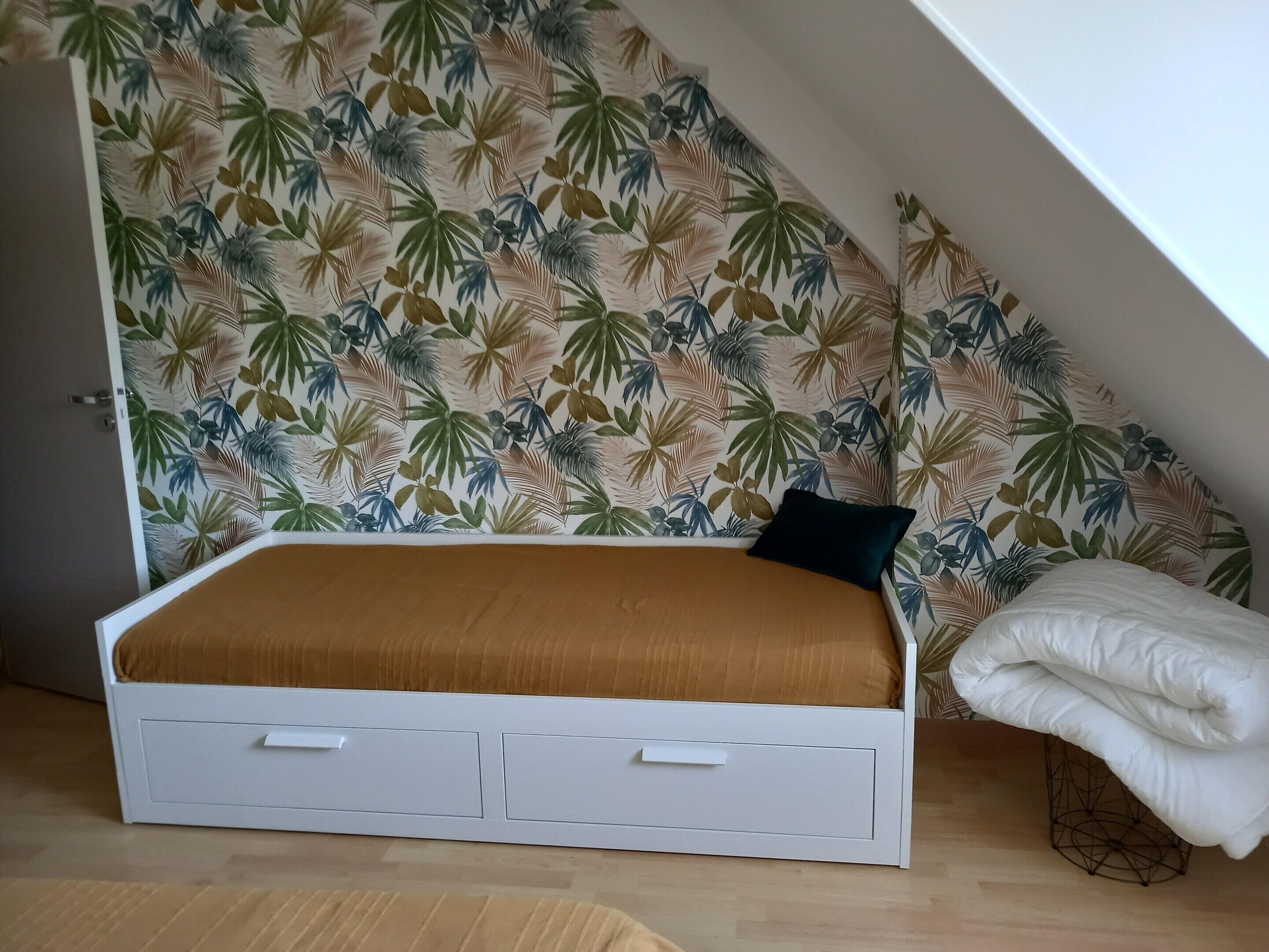 A small, white, single-bed room with a sloped ceiling, a tropical leaf-patterned wallpaper, and a wooden floor.