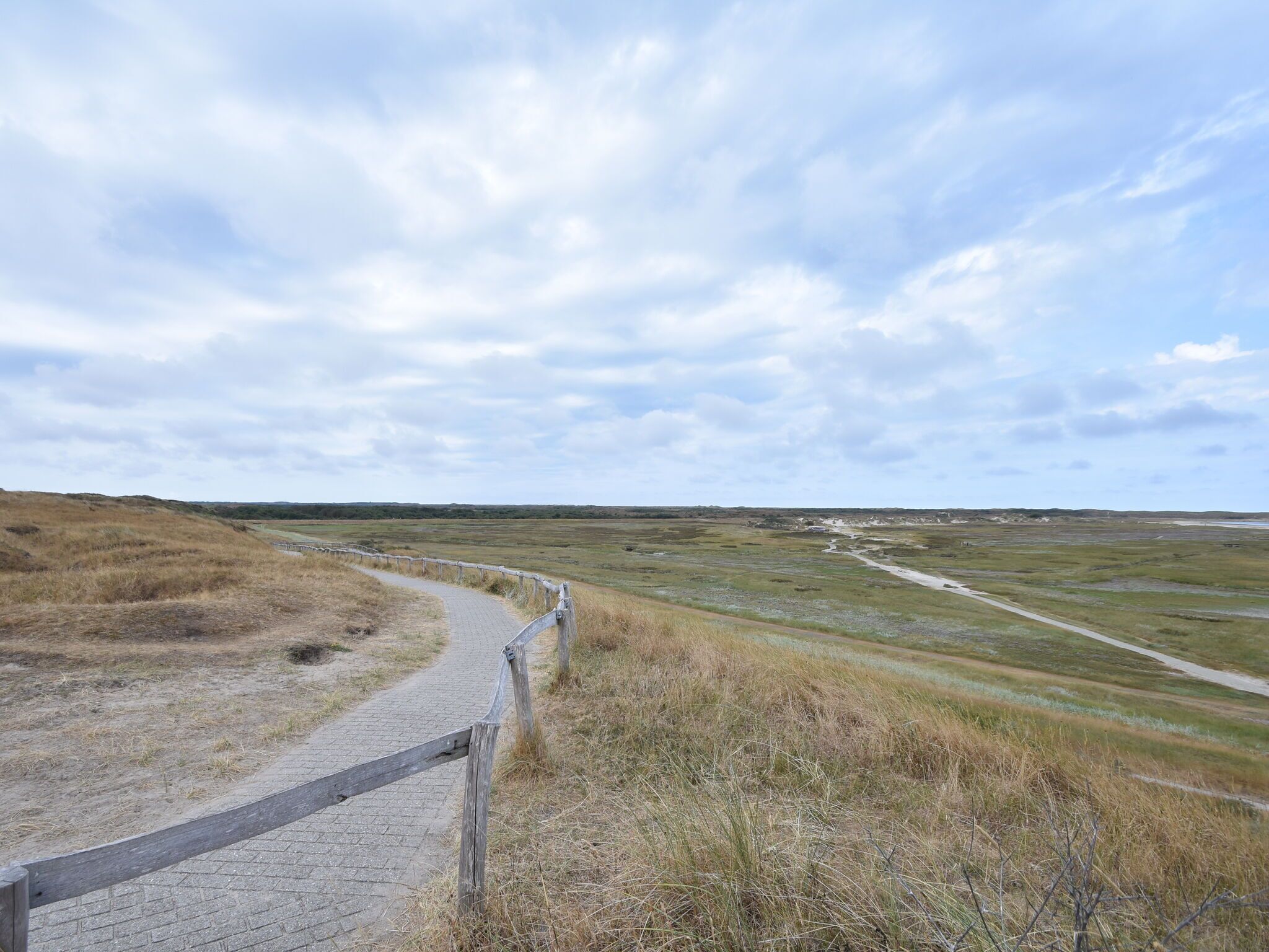 A paved pathway with a wooden fence, leading through a grassy landscape with distant hills.