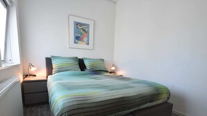 Ein ordentlich eingerichtetes Schlafzimmer mit einem Bett, zwei Nachttischlampen, einem Heizkörper und einem gerahmten Bild an der Wand.