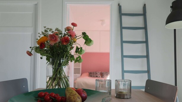 A dining table with a vase of flowers, a plate with fruit, and a ladder against the wall.