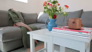 A living room with a grey sofa, a white coffee table with books, a blue vase with flowers, and a wooden mug.