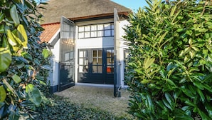 Une maison avec un toit de chaume, une porte noire et une entrée pavée, le tout entouré de verdure.