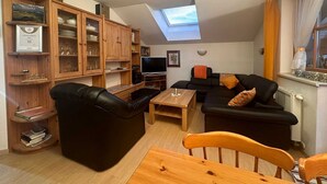 A cozy living room with a black leather sofa, wooden coffee table, and a TV. There is a wooden cabinet with glass doors and a skylight above.