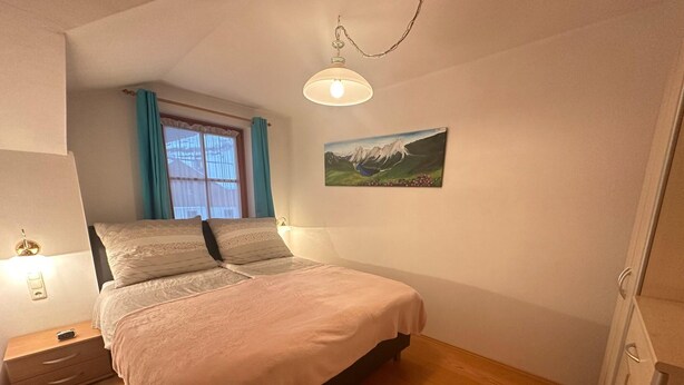 A bedroom with a bed, a nightstand, a lamp, a window with curtains, and a painting on the wall. - Bright Holiday Home in Huttschlag near Mountains Ski Slopes (Huettschlag)