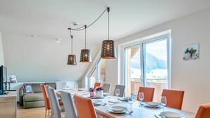 A dining area with a wooden table set for six, surrounded by orange chairs. There are pendant lights above the table, and a large window provides a view of the outdoors. - Apartment in Gosau With a Shared Sauna (Gosau)