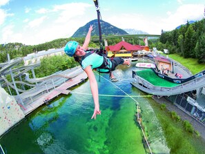 Egy személy bungee jumpingol egy magas platformról, amely egy víztömeg fölé nyúlik, a háttérben pedig egy hegy és egy épület látható.